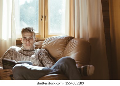 Young Man In Warm Sweater Reading Book While Relaxing On Armchair By The Window Inside Cozy Log Cabin