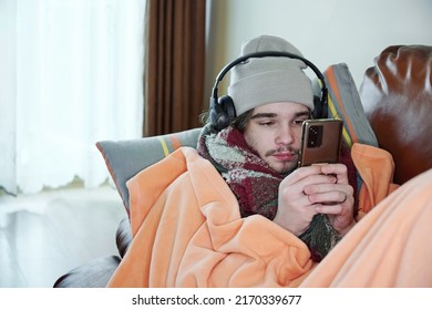 Young man warmly dressed is feeling cold, covered with warm blanket, sitting on the couch at cold home. Have problem with health, central heating