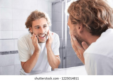 Young Man Washing Face With Soap Near Mirror In Bathroom