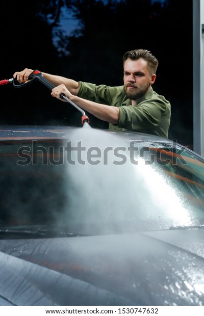 Young man washing his car in\
car wash. Cleaning Car Using High Pressure Water. Washing with\
soap.
