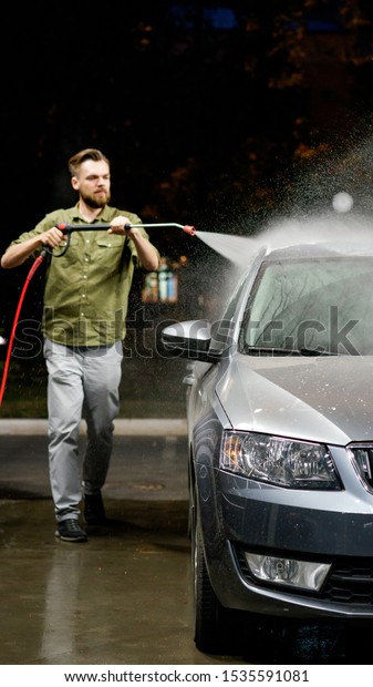 Young man washing his car in\
car wash. Cleaning Car Using High Pressure Water. Washing with\
soap.