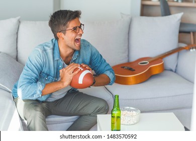 Young Man Watching American Football Game On Television Celebrating. Crazy Happy Jumping On Sofa Couch At Home With Ball Beer Bottle And Popcorn Looking Excited And Cheerful