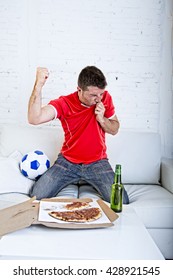young man watching football game on television kissing team jersey shield celebrating goal crazy happy jumping on sofa couch at home with ball and beer bottle with pizza excited