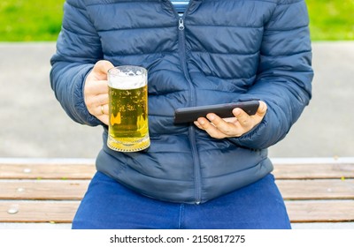 young man is watching football at the mobile phone and holding a glass of beer in another hand, in park, green nature. football fan, world cup. soccer, idols, on the go. football fanaticism, favorite 