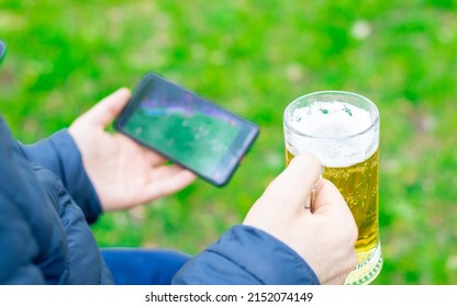 young man is watching football at the mobile phone and holding a glass of beer in another hand, in park, green nature. football fan, world cup. soccer, idols, on the go. football fanaticism, favorite 