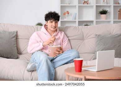 Young man watching movie with laptop and eating popcorn on sofa in living room - Powered by Shutterstock