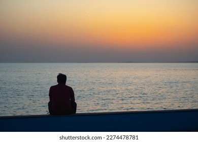 Young man watching the sea at sunset. man sitting on a wall by the ocean. Embracing the beauty and power of the ocean at sunset. - Powered by Shutterstock