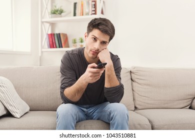 Young Man Watching Television, Using Remote Control To Switch Channels. Guy Bored With What He Sees On TV Screen. Sitting On Couch In Living Room At Home, Copy Space