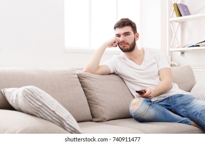 Young Man Watching Television, Using Remote Control To Switch Channels. Guy Bored With What He Sees On TV Screen. Sitting On Couch In Living Room At Home, Copy Space