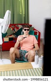 Young Man Watching TV With Remote Control In Hand, With Bathing Suit. Near The Fan. Staying At Home On Summer Vacation.