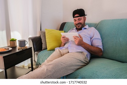 Young Man Is Watching Videos With Tablet And Sitting On The Couch - Italian Man Laughing, Sitting On The Couch And Having A Healthy Snack - Home Activities And Technology In Time Of Coronvirus 