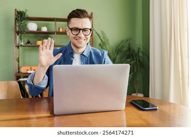 Young man waves while using a laptop in a cozy home office with green walls in the daytime - Powered by Shutterstock