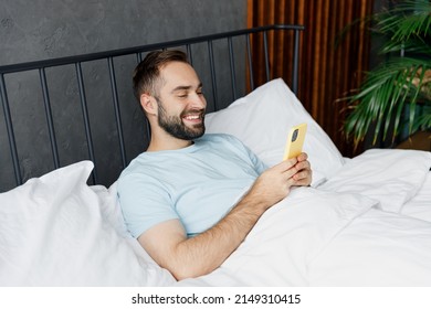 Young Man Wear Blue T-shirt Hold Use Mobile Cell Phone Chatting Online Surfing Internet Lying In Bed Rest Relax Spend Time In Bedroom Lounge Home In Own Room House Wake Up Dream. Real Estate Concept