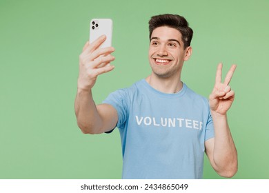 Young man wear blue t-shirt white title volunteer do selfie shot on mobile cell phone show v-sign isolated on plain pastel green background. Voluntary free work assistance help charity grace concept