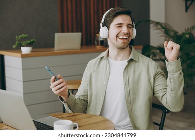 Young man wear casual clothes headphones sits alone at table in coffee shop cafe restaurant indoors work or study on laptop pc computer use cell phone dancing. Freelance mobile office business concept