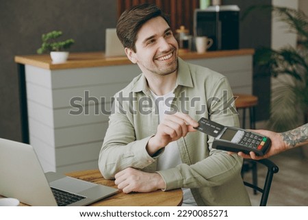 Young man wear casual clothes sitting alone at table in coffee shop cafe indoors work laptop pc computer hold bank payment terminal process acquire credit card Freelance mobile office business concept