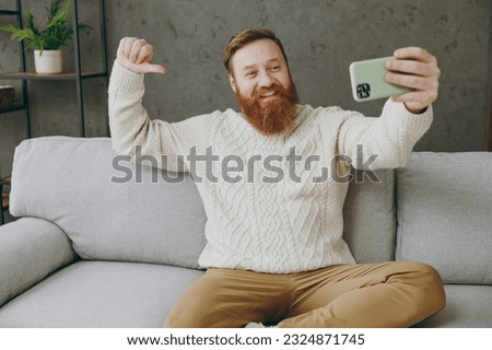 Young man wear casual clothes do selfie shot on mobile cell phone point on himself sits on grey sofa couch stay at home hotel flat rest relax spend free spare time in living room indoor Lounge concept