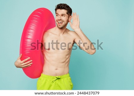 Young man wear green shorts swimsuit relax near hotel pool hold inflatable rubber ring try hear you overhear listen intently isolated on plain blue background. Summer vacation sea rest sun tan concept