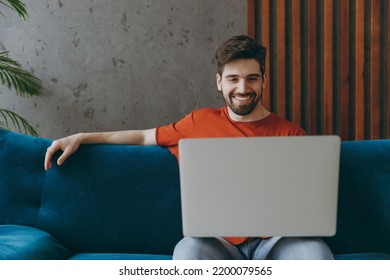 Young IT Man Wear Red T-shirt Hold Use Work On Laptop Pc Computer Sit On Blue Sofa Couch Stay At Home Hotel Flat Rest Relax Spend Free Spare Time In Living Room Indoors Grey Wall People Lounge Concept