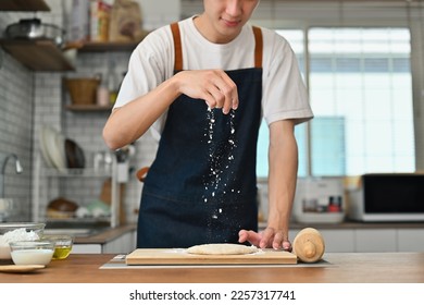Young man wearing apron sprinkling flour over fresh dough while prepared homemade pastry on kitchen table - Powered by Shutterstock