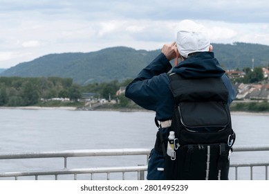 A young man wearing a backpack and cap looking through a monocular toward a village by a river. Houses line the riverbank against rolling hills under a cloudy sky, suggesting practical field observation in a rural European setting