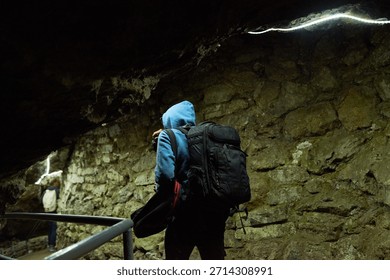 Young man wearing backpack exploring underground cave, walking along stone wall with metal railing, another middle aged Caucasian man standing in background observing surroundings