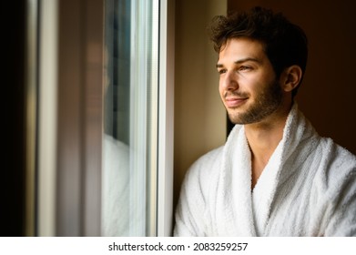 Young Man Wearing A Bathrobe And Looking Outside A Window In His Hotel Room