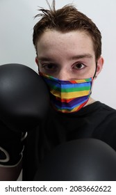 A Young Man Is Wearing Black Boxing Gloves And A Colorful Medical Mask, Standing Against A White Background. Male Youth Participates In A Sport While Staying Safe From Covid 19. Selective Focus. 