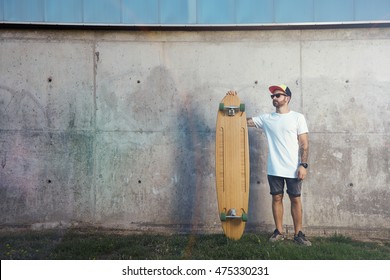 Young man wearing blank white t-shirt, baseball hat, shorts and sneakers standing with a wooden longboard next to a gray concrete wall