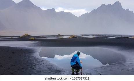 A young man wearing a blue jacket squatting on a black sand beach, in front of a paddle reflecting the mountains in a shallow sea shore. Magical and mysterious hidden gem. Beauty of the nature - Powered by Shutterstock