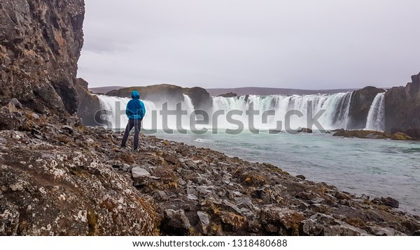 A\
young man wearing blue jacket, standing on the rocks on the side of\
the great and wide waterfall. Godafoss is called the waterfall of\
the gods. Overcast. Waterfall divided into two\
parts.\
