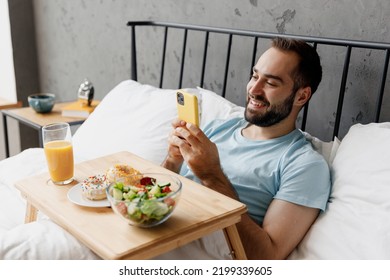 Young Man Wearing Blue T-shirt Lying In Bed Have Breakfast Taking Picture Shot On Mobile Cell Phone Of Food Rest Relax Spend Time In Bedroom Lounge Home In Own Room House. Good Mood Bedtime Concept.