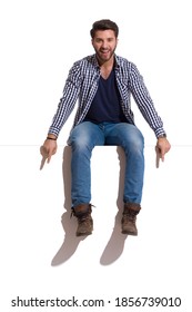 Young man wearing boots, jeans and lumberjack shirt is sitting on a top, pointing down and talkng. Full length studio shot isolated on white.