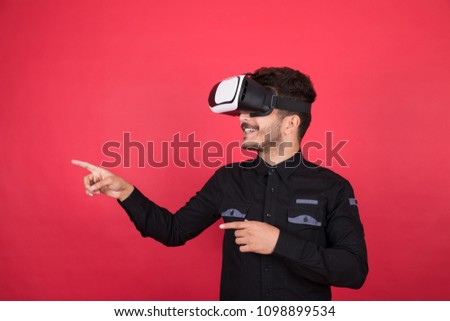 Young man wearing a casual outfit, wearing a VR glasses, looking at the right side and pointing by his fingers, standing on a red background