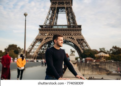 A young man wearing a dark blue jacket is standing on the background of the Eiffel Tower. Sunny weather is autumn