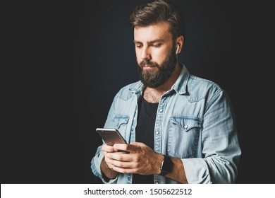 Young Man Wearing Denim Shirt And Using Smartphone. Dark-haired Bearded Hipster With Wireless Earphones Sliding Screen Of His Mobile Phone. Person In Dark Background Wearing Earpieces, Enjoying Sound.