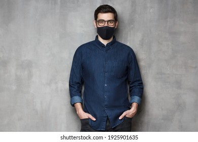 Young Man Wearing Denim Shirt, Glasses And Black Mask, Standing Against Gray Textured Wall