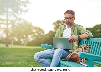 Young man wearing eyeglasses looking focused while working using laptop, sitting on the bench in the green park - Powered by Shutterstock