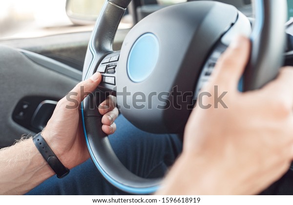 Young man wearing fitness bracelet traveling by\
new electric car transportation sitting inside holding steering\
wheel driving close-up