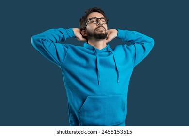 A young man wearing glasses and a casual blue hoodie stands with his hands behind his head, eyes closed in a gesture of relaxation and contentment, blue background - Powered by Shutterstock