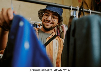 Young man wearing glasses with casual clothing and hat browsing clothes in thrift store while smiling cheerfully, engaged in the joy of shopping for unique apparel - Powered by Shutterstock