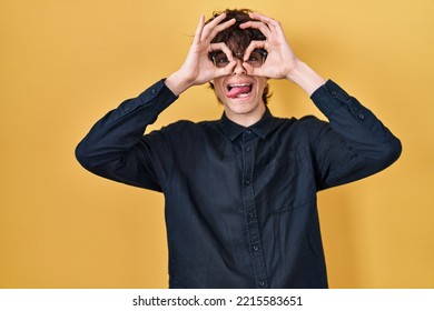 Young Man Wearing Glasses Over Yellow Background Doing Ok Gesture Like Binoculars Sticking Tongue Out, Eyes Looking Through Fingers. Crazy Expression. 