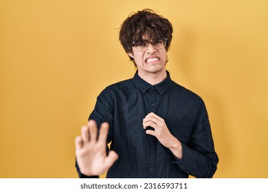 Young man wearing glasses over yellow background disgusted expression, displeased and fearful doing disgust face because aversion reaction. with hands raised 