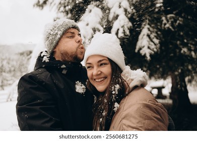 A young man wearing a gray hat covered with snow blows snow off the white hat of his smiling wife. In the background are large fir trees in the snow. Happy winter journey. - Powered by Shutterstock