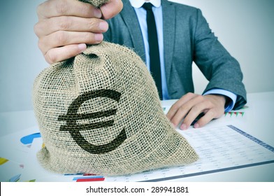 a young man wearing a gray suit seated at an office desk full of charts and financial balances holds a burlap money bag with the euro currency sign in his hand