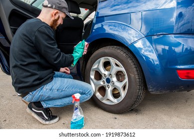 Young man wearing a green rubber glove and a cap cleaning the outside parts of a blue car with a spray and a pink microfiber cloth. Car wash concept.