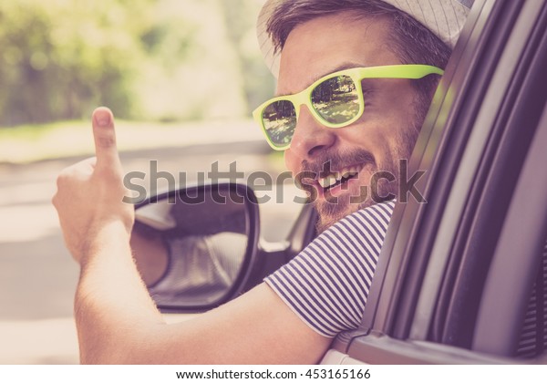 Young man wearing hat and sunglasses showing thumbs up\
from driver seat through opened window. Vacation and travel\
concepts. 