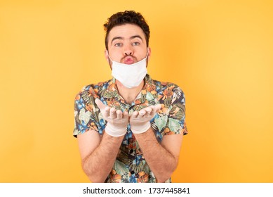 Young Man Wearing Hawaiian Shirt With Medical Mask And Gloves Against A Yellow Background Sending Blow Kiss With Pout Lips And Holding Palms To Send Air Kiss.