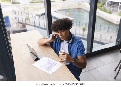 A young man wearing headphones, examining graphs on tablet. He has curly hair, holding a coffee cup with a thoughtful look, unaltered - Powered by Shutterstock