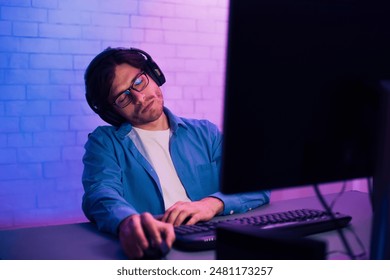 A young man wearing headphones and glasses sits in front of a computer, clearly exhausted, with his head leaning to the side. Hes working late at night, illuminated by purple and blue lights - Powered by Shutterstock